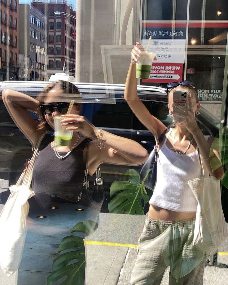 two women taking pictures in the window of a store with their cell phones and drinks