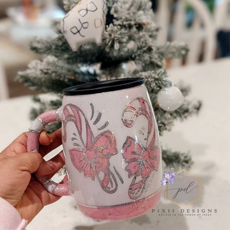 a hand holding a pink and white coffee mug in front of a small christmas tree