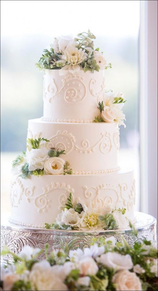 a wedding cake with white flowers and greenery on the table in front of a window