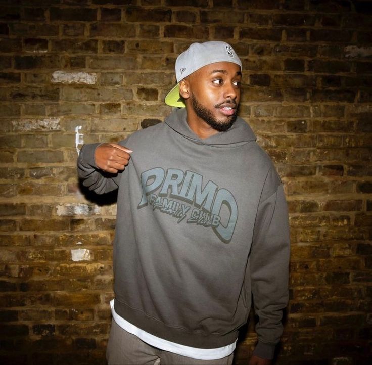 a man in a gray hoodie is standing by a brick wall and pointing to the side