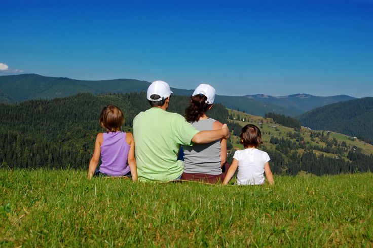 three people sitting on top of a grass covered hill looking out at the mountains and trees