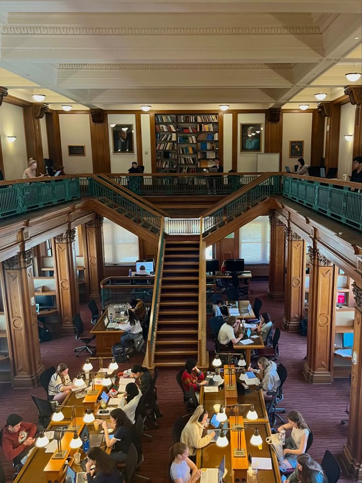 people are sitting at desks in a library with bookshelves and stairs to the second floor