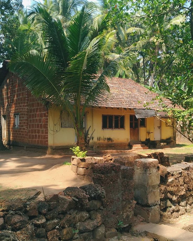 a small yellow house with a palm tree in the foreground and stone walls around it