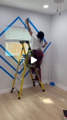 a woman on a ladder painting a wall with blue and white tape in an empty room