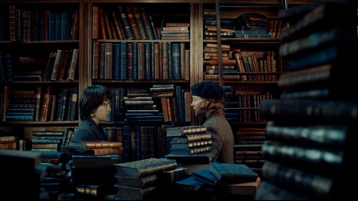 two people sitting in front of bookshelves with stacks of books on the floor