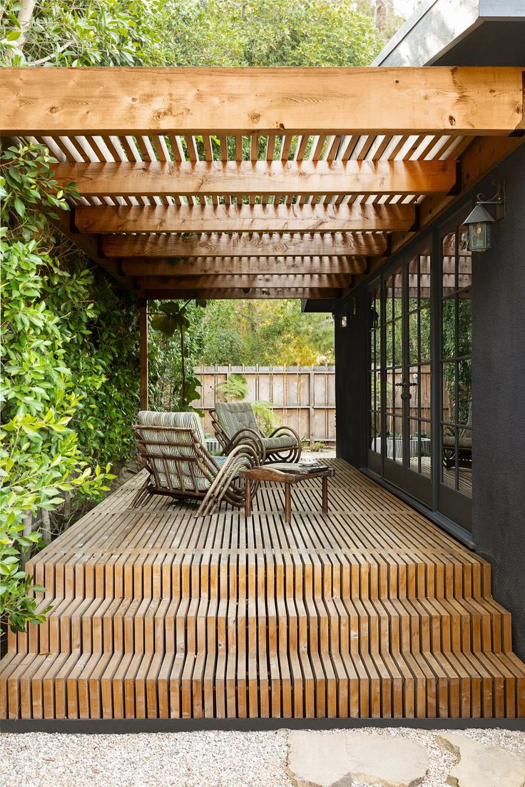 a wooden deck with two chairs under a pergolated roof and trees in the background