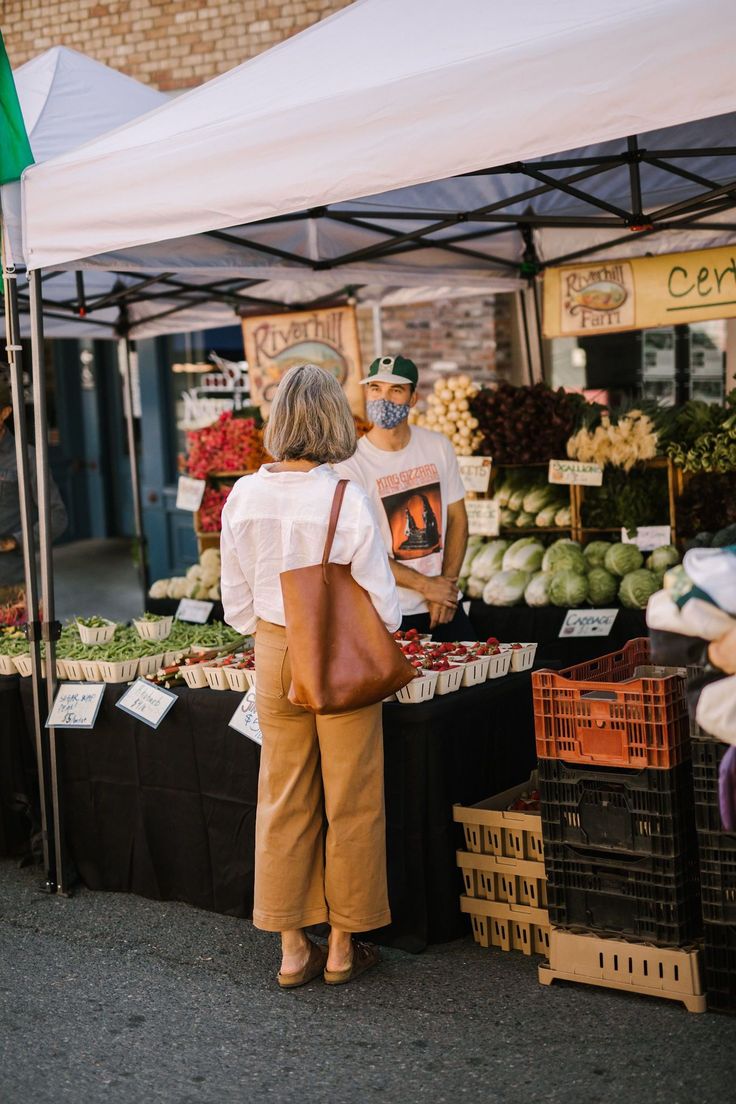 two people standing in front of a fruit and vegetable stand at an open air market