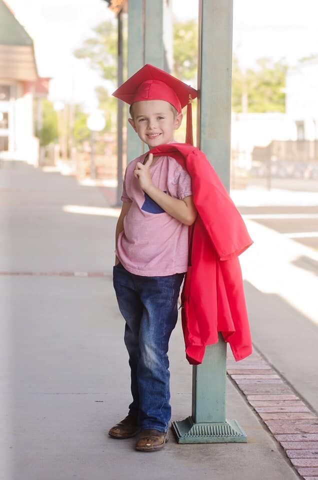 a young boy wearing a red graduation cap and gown standing next to a pole on the sidewalk