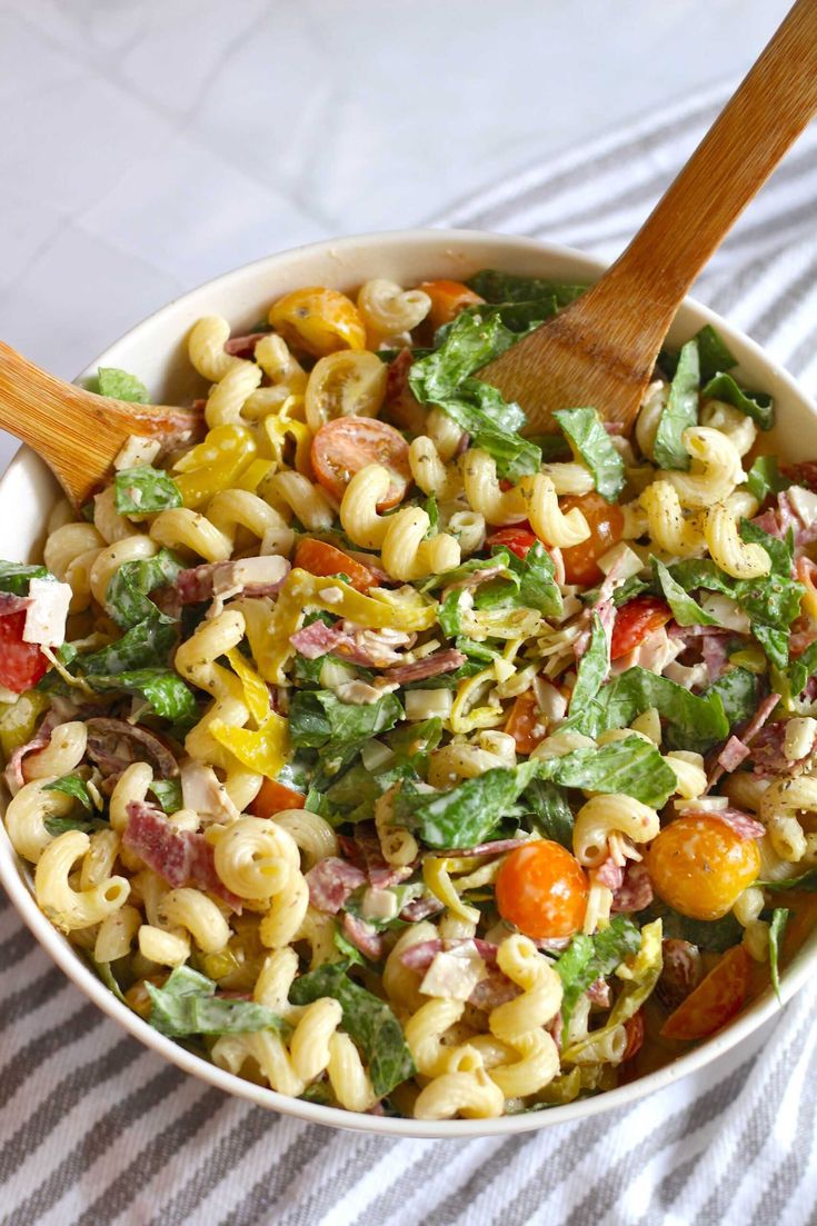 a white bowl filled with pasta salad on top of a striped table cloth next to a wooden spoon