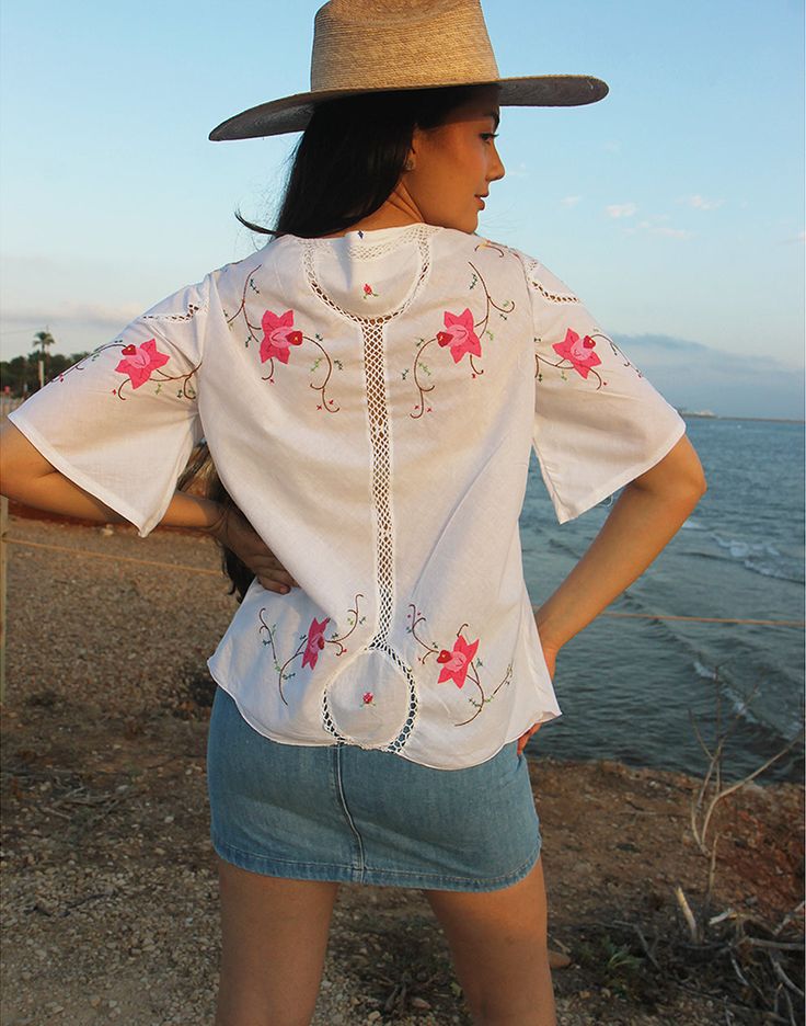 a woman wearing a white shirt with flowers on it and a hat standing next to the ocean