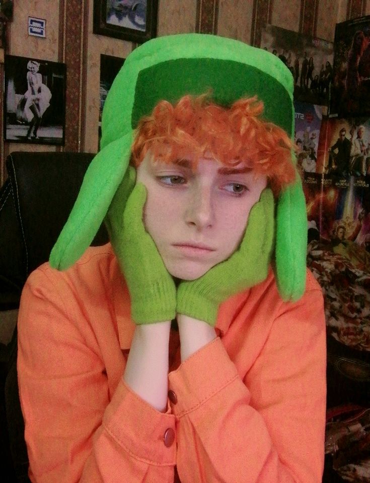 a young boy with red hair wearing a green hat and orange shirt is covering his face