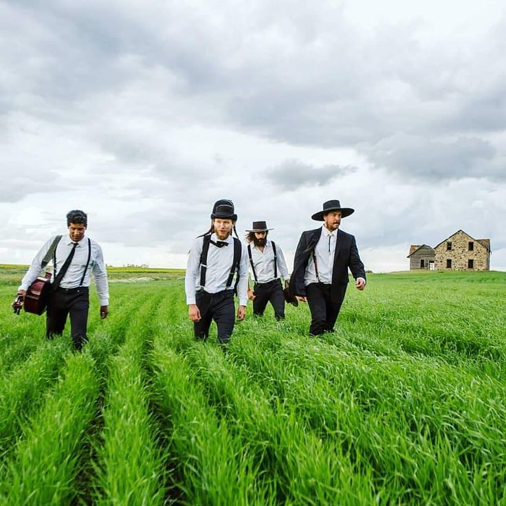 three men in suits and hats are walking through the tall grass on a cloudy day