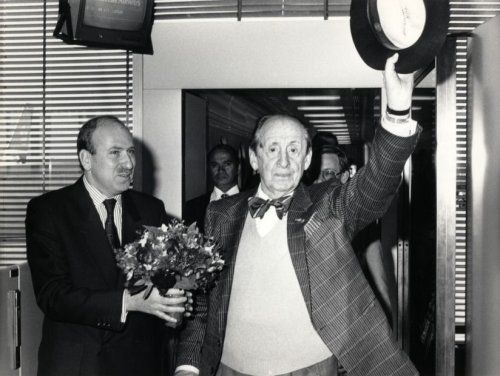 an old man in a suit and bow tie holding flowers while standing next to another man