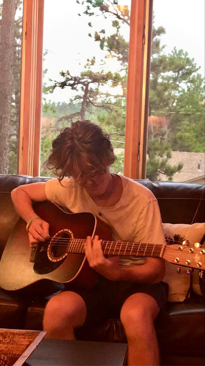 a person sitting on a couch with a guitar in front of a window playing the guitar