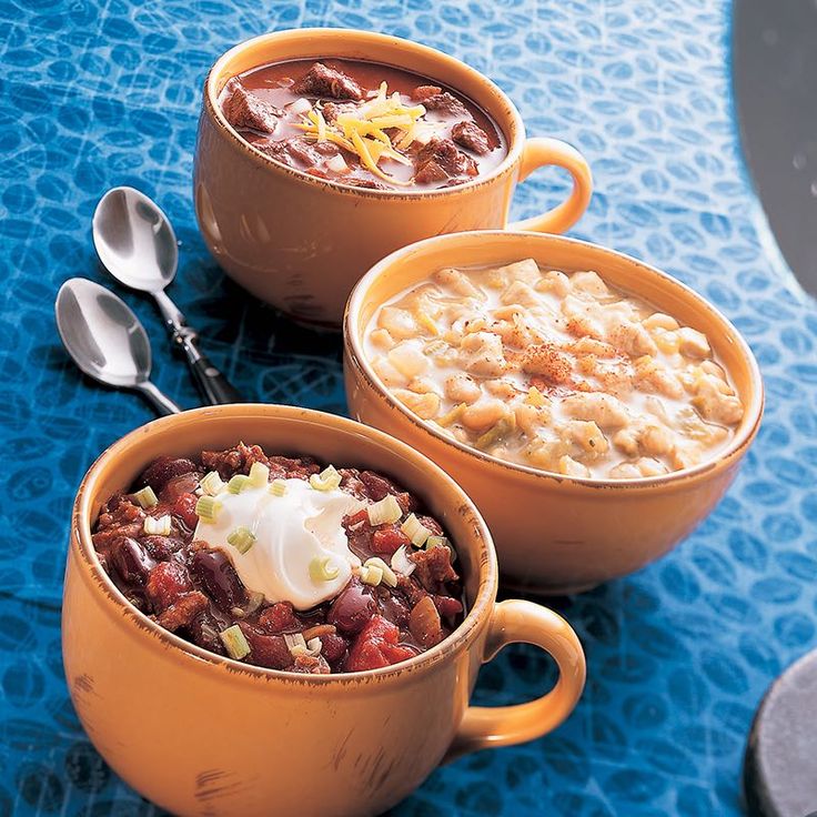 three bowls of chili and beans with spoons on a blue tablecloth next to each other