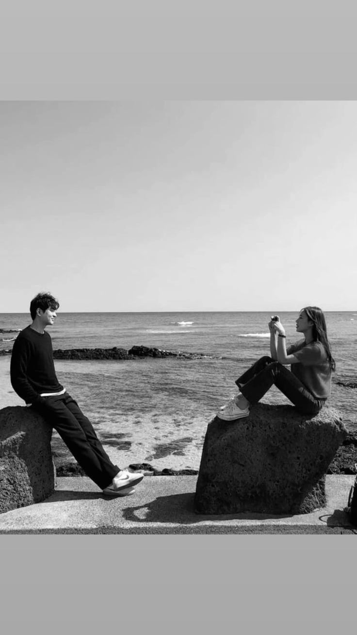 two people sitting on rocks near the ocean and one person is holding a cell phone