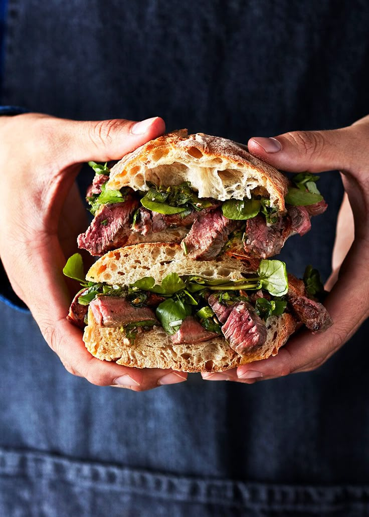 a person holding a sandwich with meat and greens in their hands, while wearing an apron