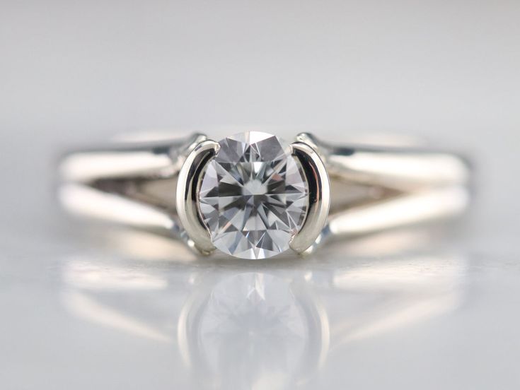 a white gold ring with a round cut diamond in the center on a reflective surface