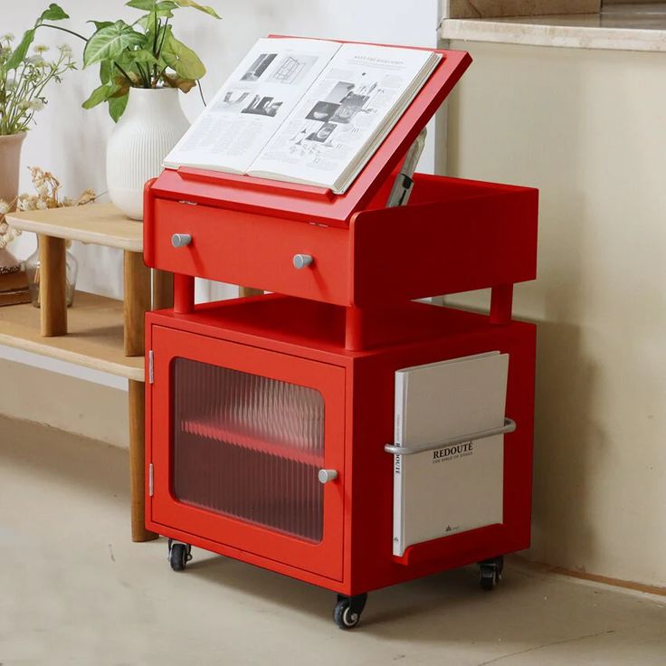 a red cart with an open magazine on top of it next to a potted plant