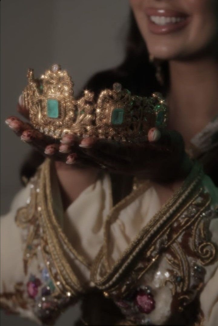 a woman holding up a gold tiara with green stones on it's sides