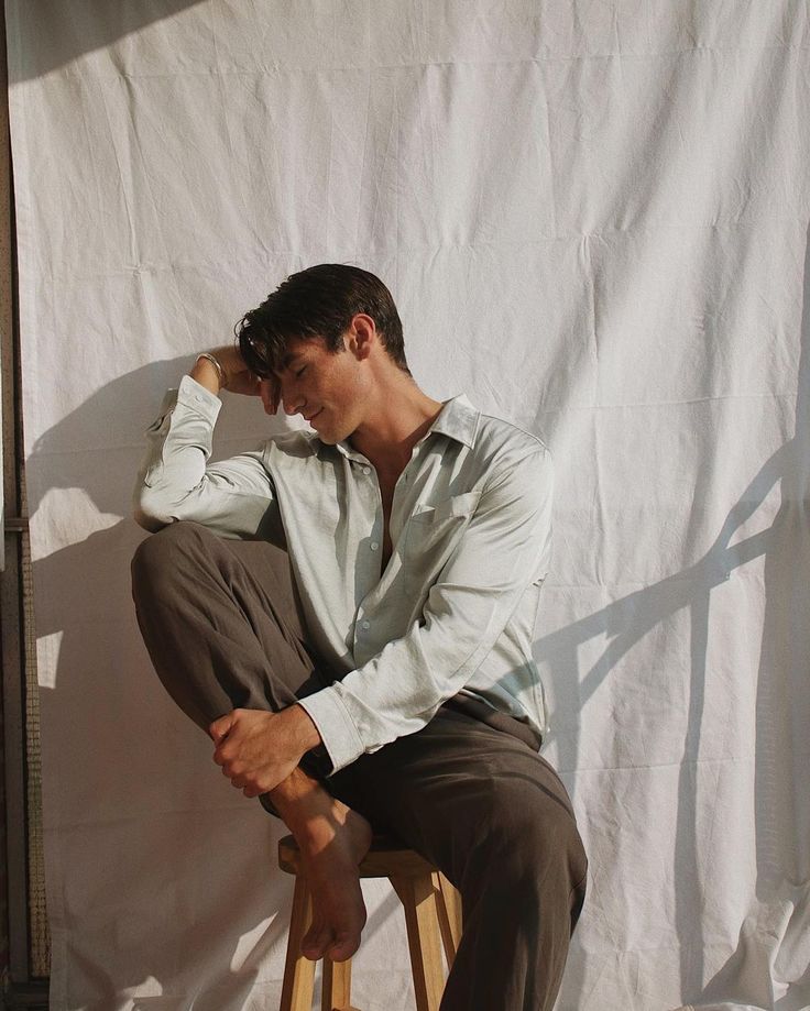 a man sitting on top of a wooden stool next to a white wall in front of a curtain