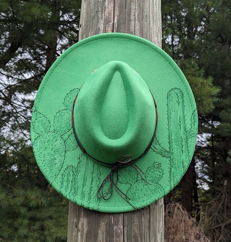 This truly one-of-a-kind fedora hat is perfect to add a special touch to your outfit or hang as a unique western wall decoration. It's burned entirely by hand using a pyrography tool with an original and entirely freehanded design (never using a stencil or laser machine.) The vivid kelly green colored material burns a deep emerald. This design features a variety of detailed cactus growing up the top of the brim. Lastly, this fedora is adorned with a brown faux leather tie back hat band that's em Cactus Burned Hat, Green Hat Bands For Kentucky Derby, Adjustable Green Wide Brim Fedora, Green Adjustable Wide Brim Fedora, Adjustable Green Wide-brim Fedora, Green Adjustable Hat Bands For Kentucky Derby, Adjustable Green Hat Bands For Kentucky Derby, Adjustable Green Fedora Felt Hat, Adjustable Wide Brim Green Felt Hat