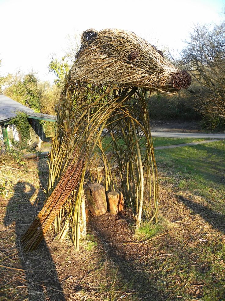 a bear sculpture made out of branches in the middle of some grass and dirt near a house