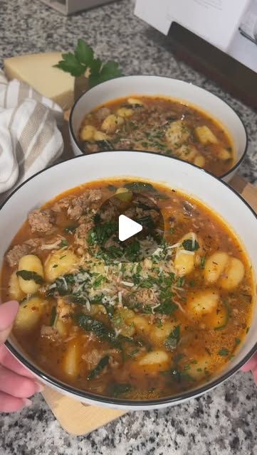 someone holding a bowl of soup in front of two bowls on a counter top with cheese and parmesan