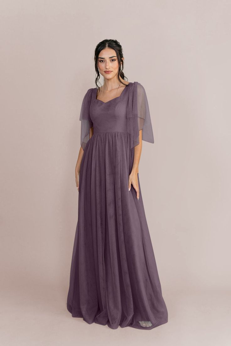 a woman in a long purple dress with sheer sleeves and an open back, standing against a