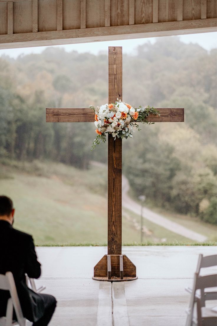 a man sitting in front of a wooden cross on top of a cement floor next to a white and orange flower arrangement