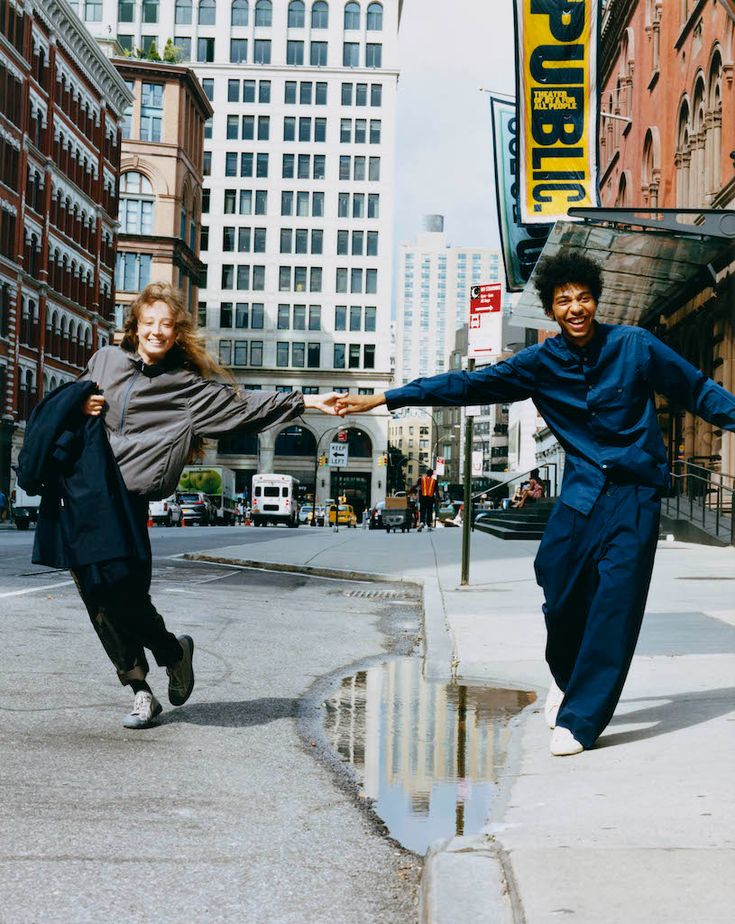 two people are dancing on the sidewalk in front of some buildings and one is holding his arms out