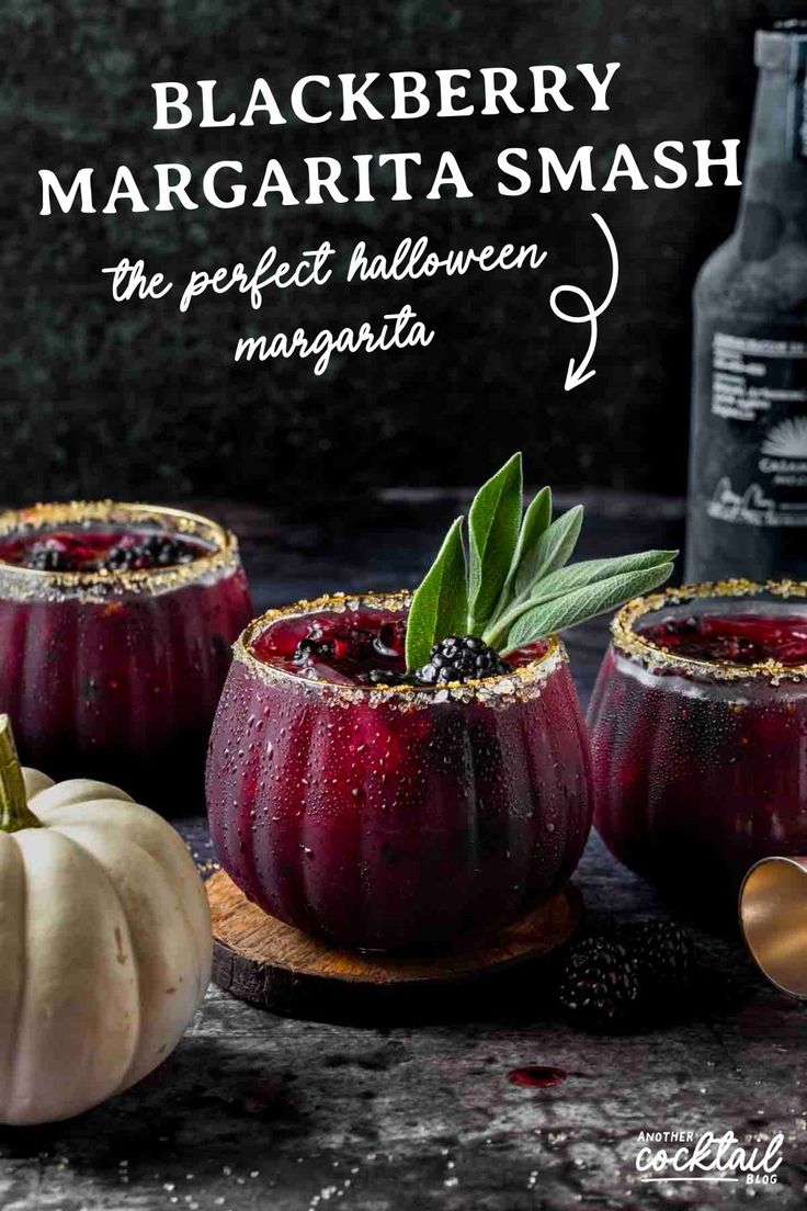 three glasses filled with black berry margarita smash on top of a wooden cutting board next to two pumpkins and a bottle of booze