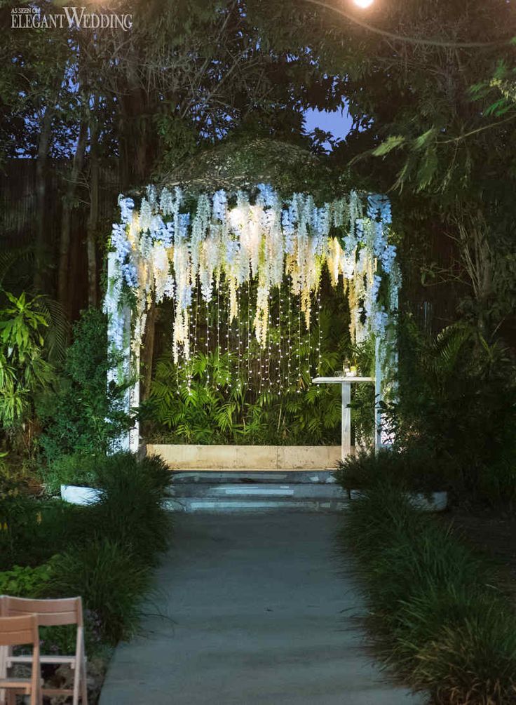 an outdoor wedding ceremony setup with white flowers and greenery on the wall, surrounded by string lights