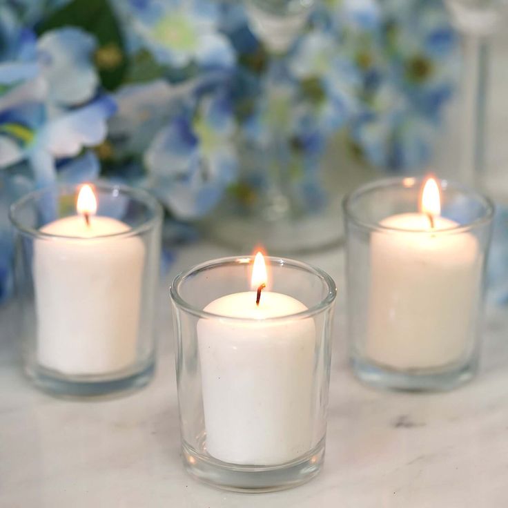 three white candles sitting on top of a table next to flowers and vases with blue hydrangeas in the background