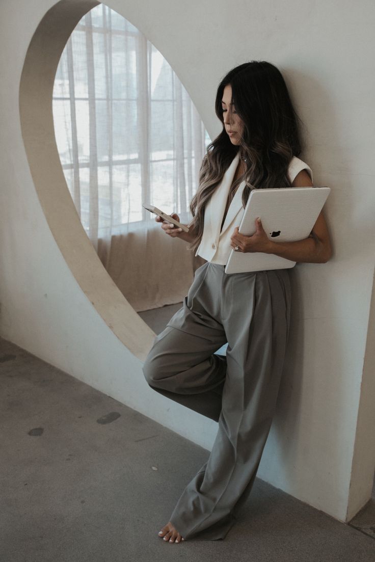 a woman leaning up against a wall using her cell phone and laptop while holding a tablet