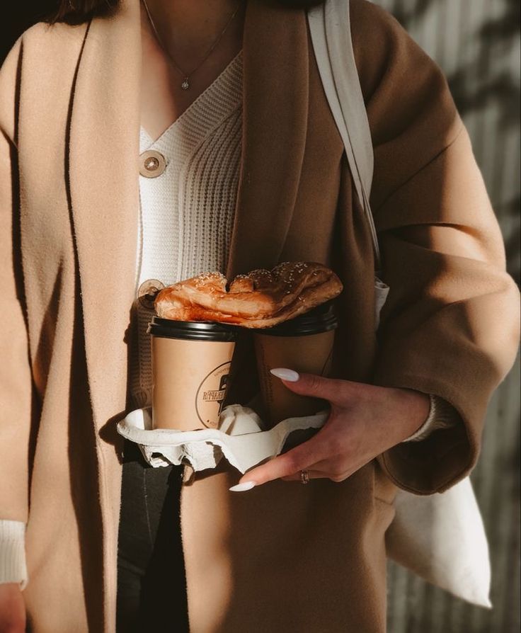 a woman holding a coffee cup and food item in her hand while wearing a brown coat