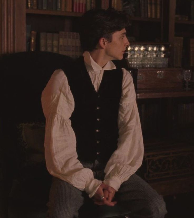 a man sitting on a chair in front of a bookshelf wearing a vest