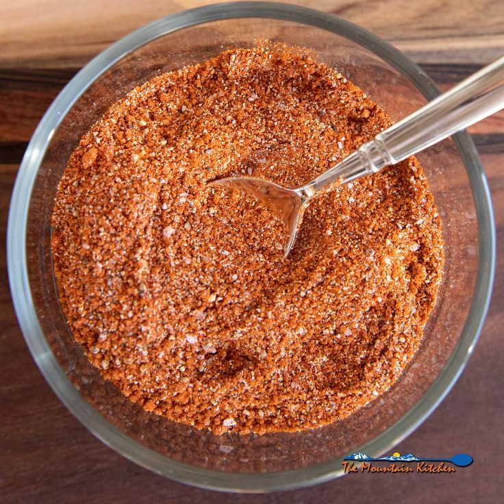 a glass bowl filled with spices on top of a wooden table next to a spoon