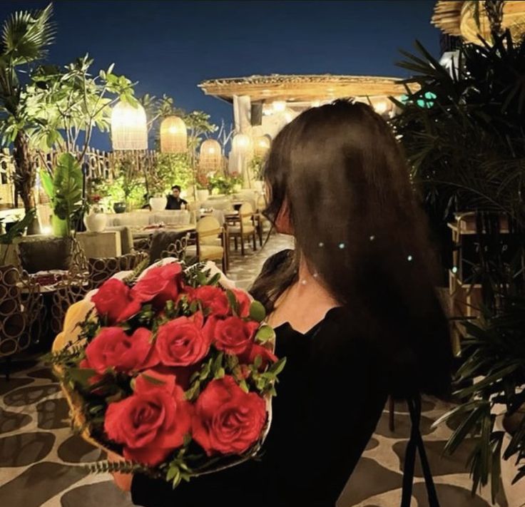 a woman holding a bouquet of roses in front of a restaurant at night with the lights on