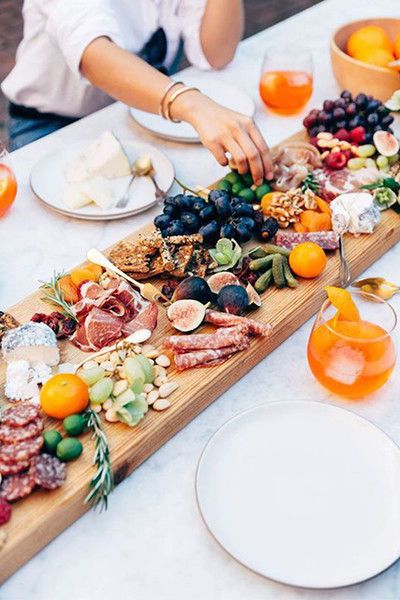 two people are serving food on a long wooden platter with oranges and grapes