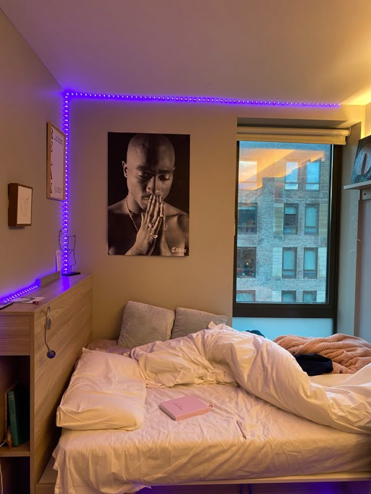 an unmade bed in front of a window with blue lights on the side and a poster above it