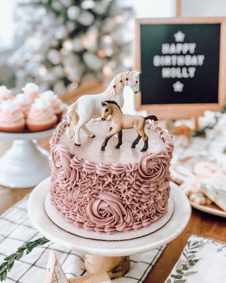 a cake with pink frosting and two horses on top, sitting on a table next to cupcakes