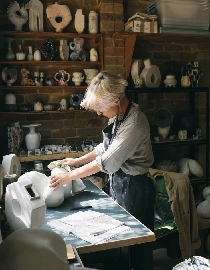 an older woman is working on pottery in her studio with lots of shelves and vases behind her