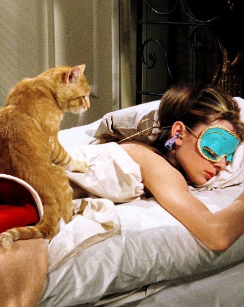 a woman laying in bed with a cat on her head and sleeping mask over her eyes