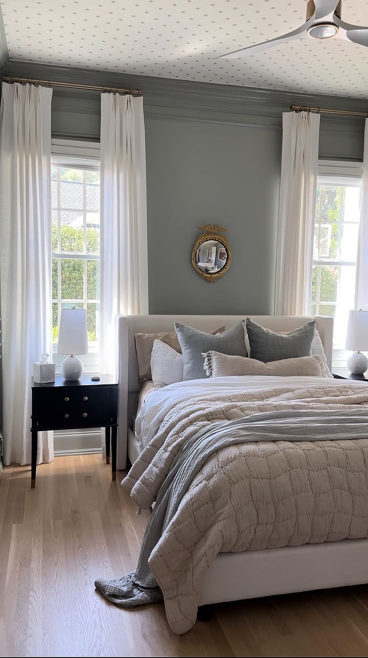 a bed sitting in a bedroom next to two windows with white drapes on them