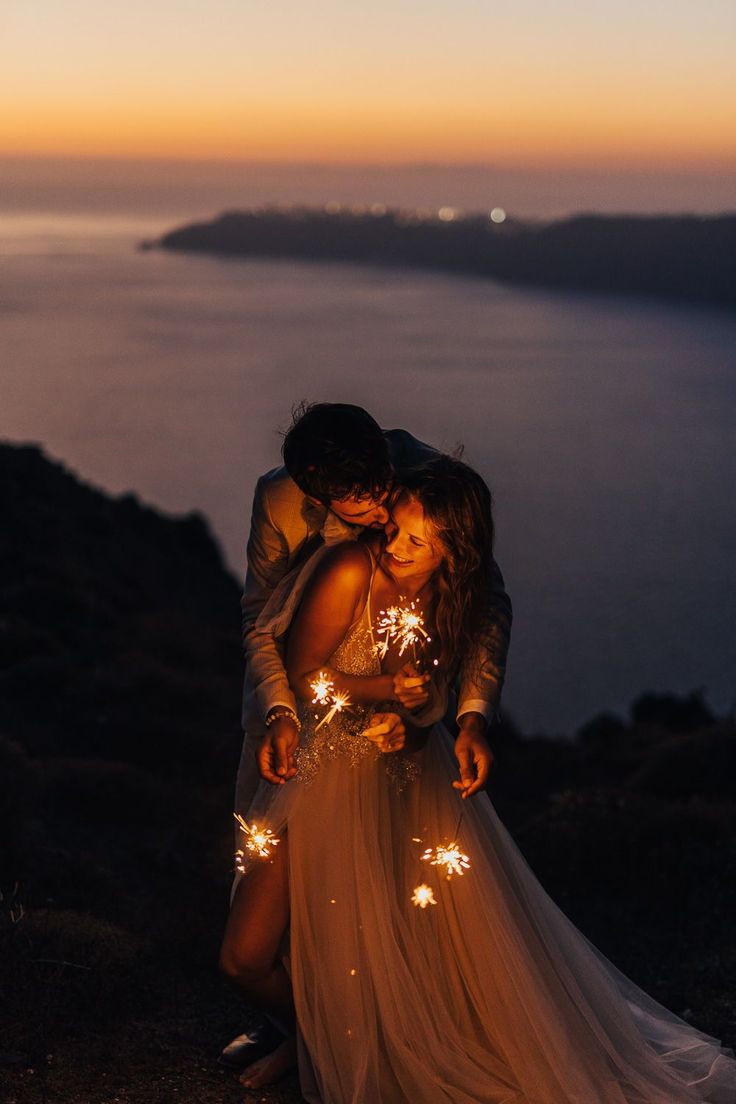 a man and woman standing next to each other holding sparklers in their hands while the sun is setting