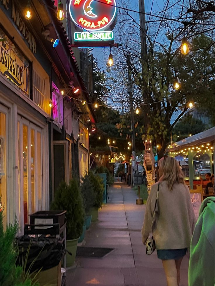 a woman walking down the sidewalk in front of a restaurant with lights hanging above it