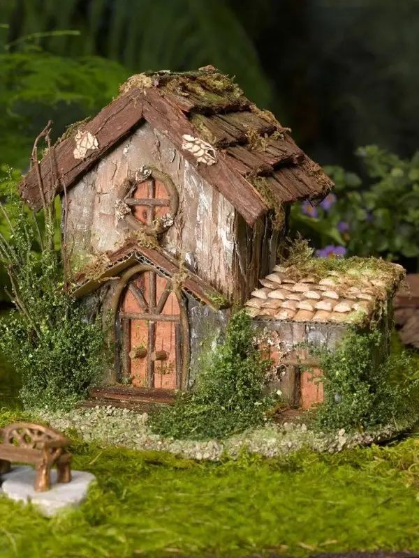 a miniature house made out of wood and stone with moss growing on the roof, along with a bench in front of it