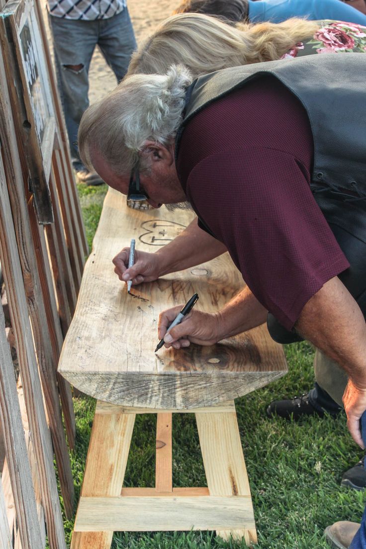 an older man writing on a piece of wood while standing next to other people in the grass