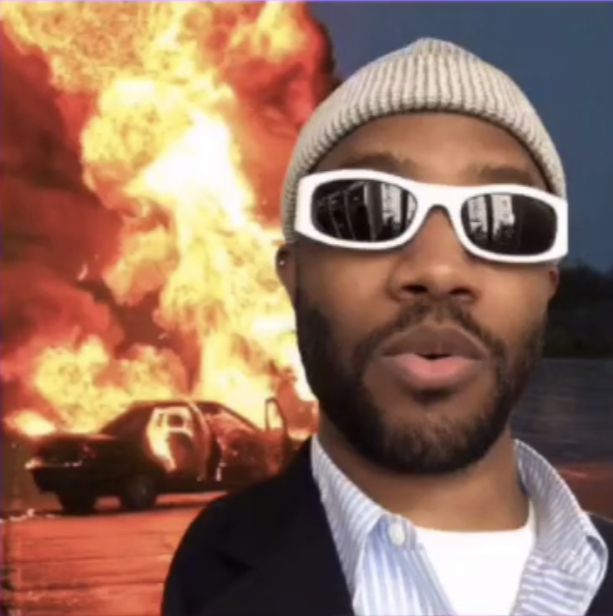 a man wearing sunglasses and a hat standing in front of a large fire with cars on it